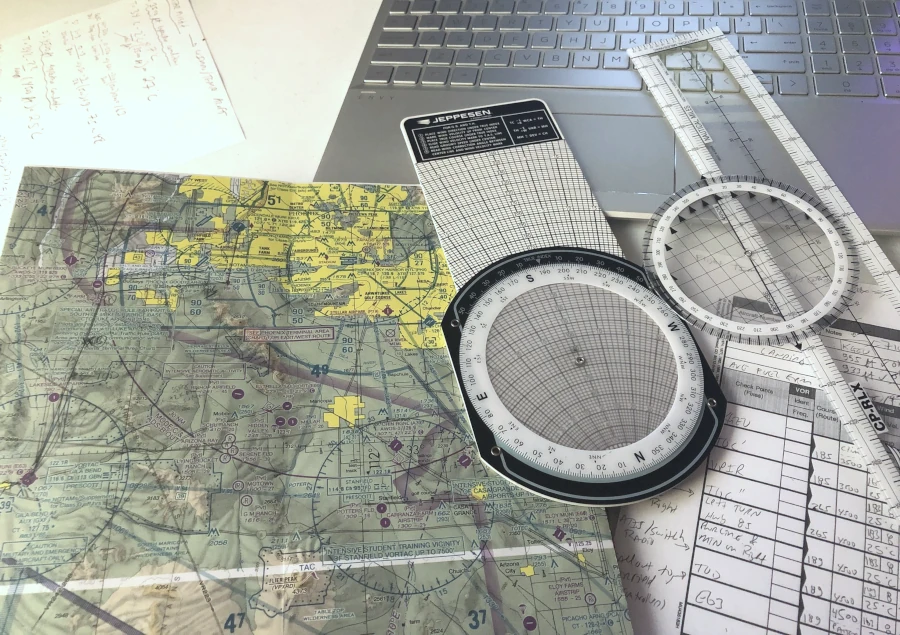 Phoenix-area sectional chart with an E6B flight computer, a plotter, and a handwritten navigation log on a desk